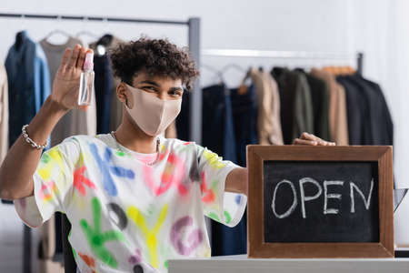 African american owner of showroom in protective mask holding hand sanitizer and chalkboard with...