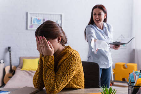 teenage girl covering face with hands near angry mother tearing papers in notebookの写真素材
