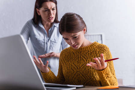 blurred mother arguing with dissatisfied teenage girl near laptop