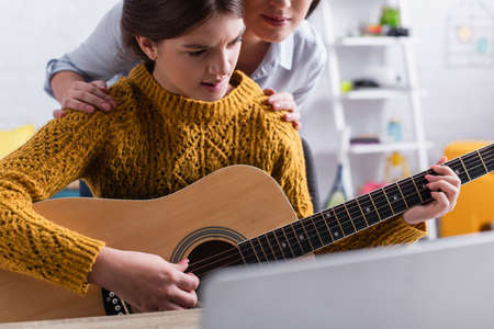 teenager biting lips while learning how to play acoustic guitar near mother on blurred backgroundの写真素材