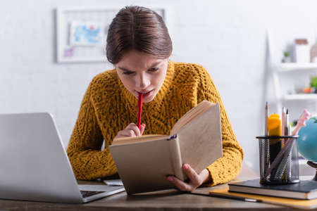thoughtful teenage girl reading book and holding pen while studying online