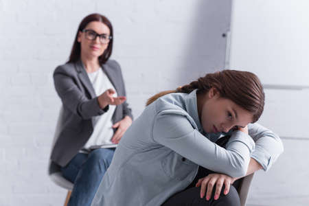 frustrated teenage girl looking away with psychologist sitting behind on blurred backgroundの写真素材