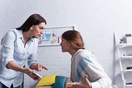 mother pointing with finger at smartphone while quarrelling with offended daughterの写真素材