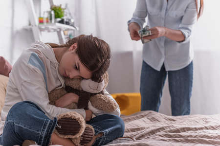 upset teenage girl hugging teddy bear near mother on blurred backgroundの写真素材
