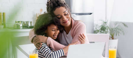 happy african american mom and daughter embracing while watching film on laptop, blurred foreground,...
