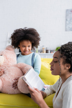 african american woman reading book to daughter sitting on sofa with teddy bearの写真素材