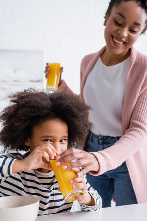 african american kid drinking orange juice from glass in hand of mother on blurred backgroundの写真素材