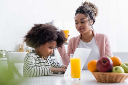 smiling african american woman looking at concentrated daughter using laptop on blurred foregroundの写真素材