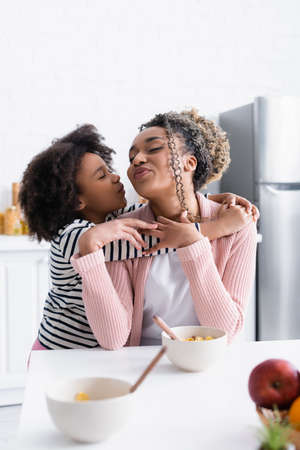 african american girl embracing and kissing mother in kitchen, blurred foreground