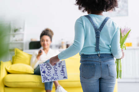 back view of african american girl with flowers and happy mothers day card near mom on blurred...