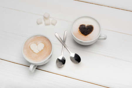 top view of cups with cappuccino and latte near spoons and sugar cubes on white wooden surfaceの写真素材
