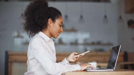 side view of smiling african american woman holding smartphone and using laptop in cafe