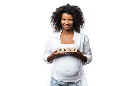Smiling pregnant african american woman holding cubes with baby lettering isolated on whiteの写真素材