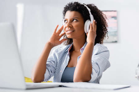 Smiling african american woman in headphones sitting near laptop on blurred foreground at home