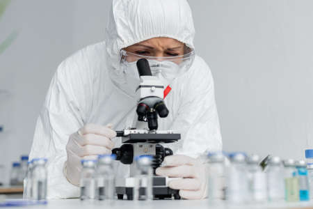 Scientist in goggles and hazmat suit looking at microscope near vaccines on blurred foregroundの写真素材