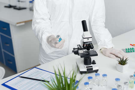 Cropped view of scientist in hazmat suit holding vaccine near microscope and clipboardの写真素材