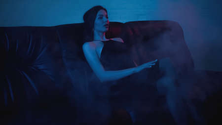 brunette woman in dress sitting on black couch on blue with smokeの写真素材