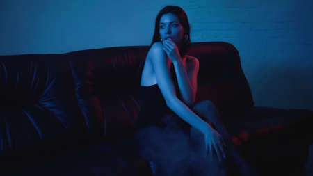 red lighting on seductive brunette woman on black couch on blue with smokeの写真素材