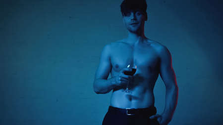 shirtless man holding glass of wine while standing with hand in pocket on blue