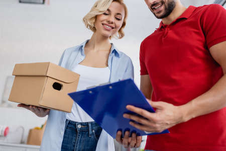 low angle view of delivery man holding clipboard near blonde woman, blurred foreground