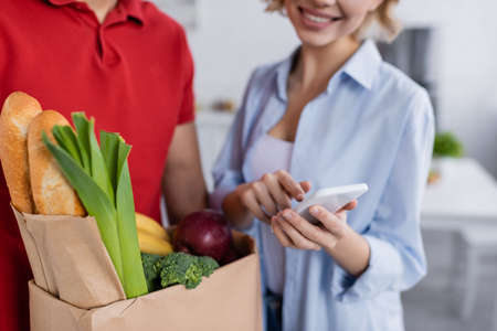 partial view of delivery man with fresh food in paper bag near happy woman pointing at smartphone on...