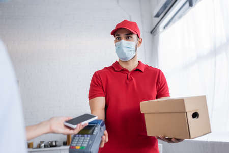 muslim courier in medical mask holding payment terminal near woman with smartphone on blurred...