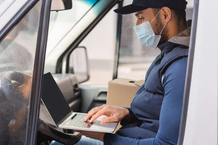 arabian delivery man in medical mask typing on laptop while sitting in car