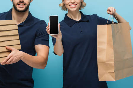 cropped view of smiling courier showing smartphone with blank screen near colleague on blueの写真素材