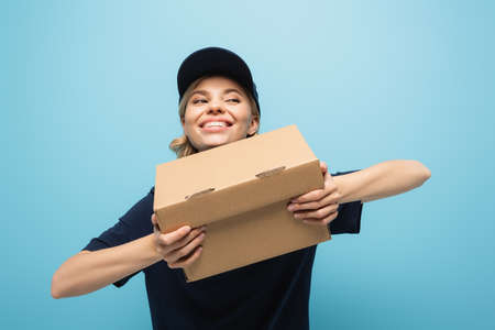 excited courier looking away while holding carton box isolated on blue