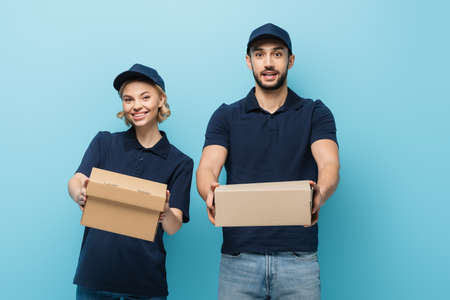 positive interracial couriers showing cardboard boxes while looking at camera isolated on blueの写真素材