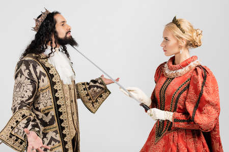 blonde queen holding sword near neck of hispanic king in medieval clothing and crown isolated on whiteの写真素材