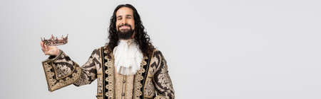 pleased hispanic king in medieval clothing holding golden crown while looking at camera isolated on white, bannerの写真素材