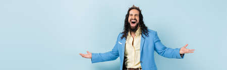 excited hispanic man in crown and jacket standing with outstretched hands isolated on blue, bannerの写真素材