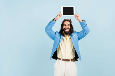 happy hispanic man in jacket and crown holding digital tablet with blank screen above head isolated...