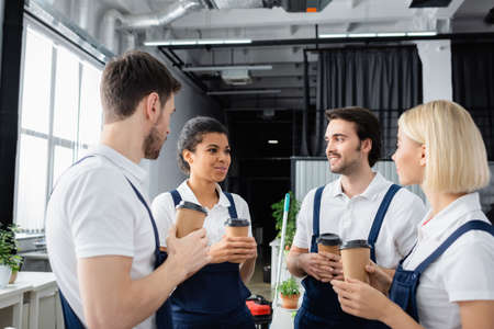 Smiling african american cleaner in overalls talking to colleagues during coffee break in officeの写真素材