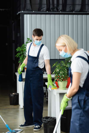 Cleaner in medical mask and uniform cleaning table near colleague on blurred foreground in officeの写真素材