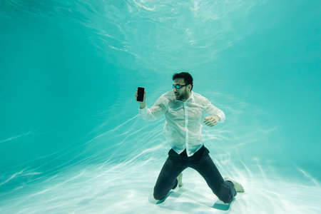 Arabian businessman in formal wear and goggles holding smartphone with blank screen underwaterの写真素材