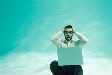 Amazed arabian businessman in goggles looking at camera near laptop in swimming pool