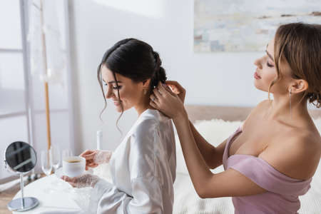 young woman making hairstyle to smiling bride holding coffee in bedroom