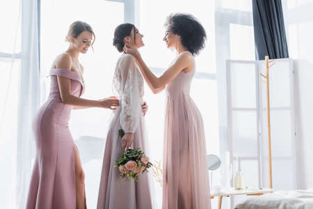 multicultural bridesmaids adjusting dress of happy bride holding wedding bouquetの写真素材