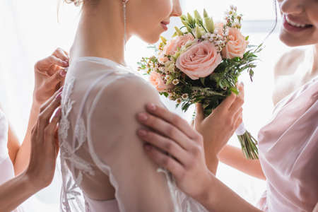 cropped view of bridesmaids preparing smiling bride for wedding, blurred foreground