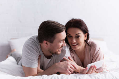 Cheerful adult couple using smartphone while lying on bedの写真素材