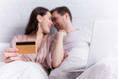 Laptop near woman holding credit card and looking at husband on blurred background on bed