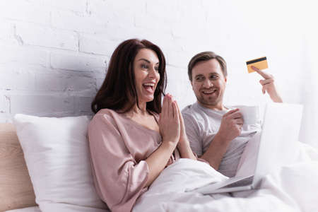 Positive woman with praying hands looking at laptop near husband with credit card and cup in bedroom