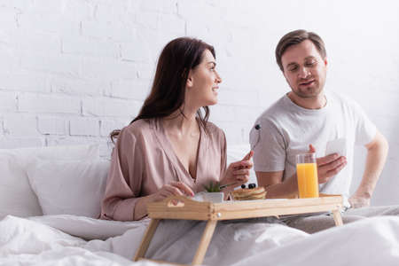 Woman looking at husband with smartphone near breakfast on blurred foreground on bedの写真素材