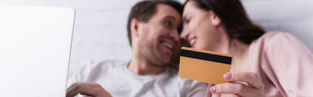 Credit card in hand of woman smiling near husband with laptop on blurred background, bannerの写真素材