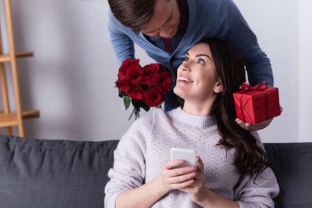 Smiling woman holding smartphone near husband with flowers and presentの写真素材