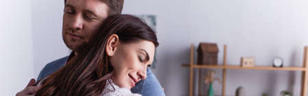 Adult man embracing wife with closed eyes at home, banner
