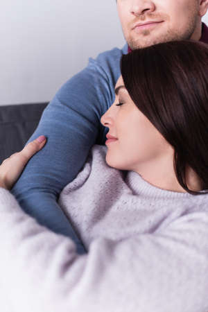 Brunette woman with closed eyes hugging husband on couchの写真素材