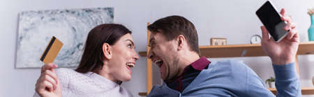 Amazed couple with credit card and smartphone looking at each other at home, bannerの写真素材